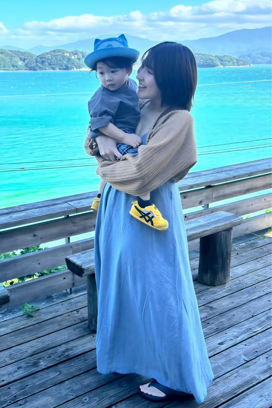 【海辺のデッキで幼い息子を抱きながら笑顔を見せるシングルマザー。自然の景色の中で穏やかな時間を過ごしている。】