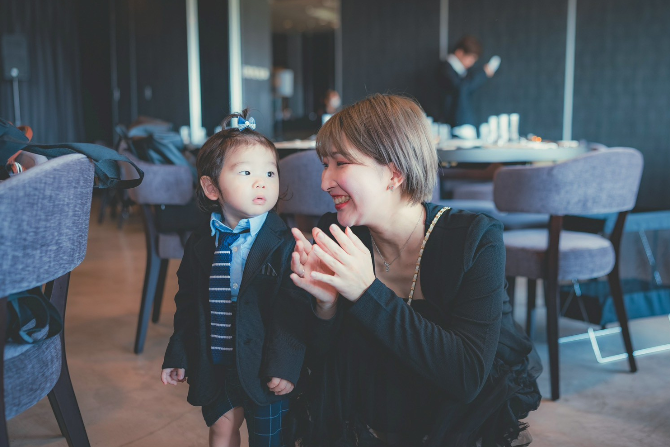 【フォーマルな服装をした幼い息子と笑顔で向き合うシングルマザー。室内で親子の温かな時間を過ごしている様子。】